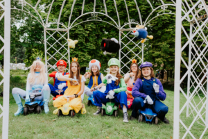 Gruppenselfie von Cosplayern in bunten Kostümen, die Charaktere aus dem Mario-Universum darstellen, während sie auf kleinen Fahrzeugen sitzen. Die Szene findet im Freien statt, umgeben von Bäumen und einem dekorativen Pavillon, passend zum Elbenwald Festival.
