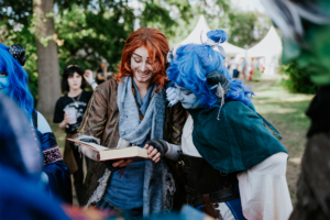 Zwei Cosplayer auf dem Elbenwald Festival, einer mit rot-orangefarbenen Haaren und der andere mit blauen Haaren und spitzen Ohren, betrachten gemeinsam ein Buch. Im Hintergrund sind weitere Festivalbesucher und Zelte sichtbar, die die Atmosphäre des Events unterstreichen.