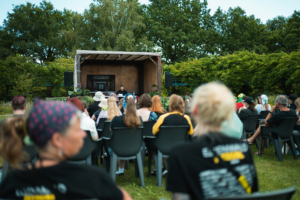Akram El-Bahay Auf dem Elbenwald Festival lauschen Besucher einem Vortrag auf einer Bühne im Freien, umgeben von einer grünen Landschaft. Die Zuschauer tragen verschiedene Outfits und sitzen in Stühlen, während ein Redner auf der Bühne spricht.