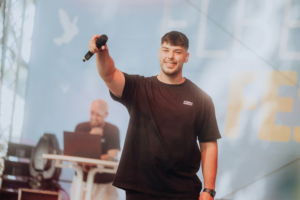 Anbu Studios Ein junger Mann steht auf der Bühne des Elbenwald Festivals, hält ein Mikrofon in der Hand und lächelt. Im Hintergrund ist ein DJ an einem Laptop zu sehen. Die Atmosphäre ist lebhaft und feierlich, typisch für ein Musik- und Nerd-Event.