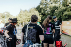Besucher des Elbenwald Festivals tragen schwarze T-Shirts mit bunten Schriftzügen, während sie sich in einer lebhaften Umgebung bewegen. Im Hintergrund sind grüne Bäume und Festivalbannern sichtbar, die die Atmosphäre des Events unterstützen.