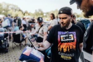 Zwei Teilnehmer des Elbenwald Festivals stehen in einer Menschenmenge, während einer auf sein Smartphone schaut. Der Mann trägt ein schwarzes T-Shirt mit dem Elbenwald Festival-Logo. Im Hintergrund sind weitere Festivalbesucher und Stühle zu sehen.