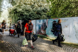 Besucher des Elbenwald Festivals tragen Gepäck und Ausrüstung auf einem gepflasterten Weg, umgeben von grüner Natur und Festival-Werbung. Die Szene zeigt die Vorfreude der Teilnehmer auf das bevorstehende Event.