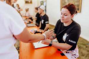 Mitarbeiterin des Elbenwald Festivals registriert einen Besucher an einem orangefarbenen Tisch. Im Hintergrund sind weitere Helfer zu sehen, die an Laptops arbeiten. Die Atmosphäre ist freundlich und einladend, passend zu einem nerdigen Musik- und Kultur-Event.