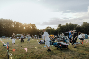 Festivalbesucher beim Elbenwald Festival, das eine Mischung aus Musikfestival und Convention bietet. Die Szene zeigt Teilnehmer, die sich mit Ausrüstung in einem Zeltlager auf einem sonnigen Feld bewegen, umgeben von bunten Fähnchen und Zelten.