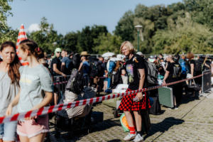 Menschenmenge beim Elbenwald Festival, mit Teilnehmern in bunten Outfits, die auf dem Gelände anstehen. Im Vordergrund ein junger Mann in einem karierten Rock und T-Shirt, umgeben von Festivalbesuchern und Gepäck, bei strahlendem Sonnenschein.