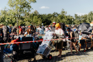Menschenmenge beim Elbenwald Festival, die mit Wagen und Gepäck ansteht. Teilnehmer tragen bunte Outfits und transportieren Zelte, Schlafsäcke und andere Festival-Utensilien unter strahlend blauem Himmel. Atmosphäre von Vorfreude und Gemeinschaft.
