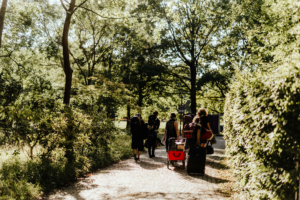 Festivalbesucher auf dem Weg zum Elbenwald Festival, umgeben von grüner Natur. Die Gruppe zieht Wagen und Gepäck auf einem gepflasterten Weg, während die Sonne durch die Bäume scheint und eine festliche Stimmung verbreitet.