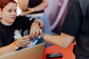 Eine Mitarbeiterin des Elbenwald Festivals befestigt ein Armband an einem Besucher, während ein Laptop und ein Smartphone auf dem Tisch liegen. Die Szene vermittelt die Atmosphäre des Festivals, das Musik und Nerd-Kultur vereint.
