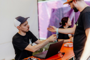 Ein Mitarbeiter des Elbenwald Festivals überprüft die Eintrittsarmbänder eines Besuchers an einem Registrierungstisch. Im Hintergrund sind weitere Teammitglieder zu sehen, die für den reibungslosen Ablauf des Events sorgen.