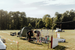 Zwei Personen bereiten ihre Zelte auf dem Elbenwald Festival vor, umgeben von einer bunten Wiese und Dekoration. Im Hintergrund sind weitere Zelte und eine festliche Atmosphäre sichtbar, die das einzigartige Erlebnis des Festivals unterstreicht.