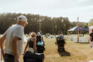 Besucher des Elbenwald Festivals ziehen mit Wagen und Gepäck über das Gelände, um ihre Zelte aufzustellen. Im Hintergrund sind Zelte und Festivaleinrichtungen unter blauem Himmel sichtbar, während die Veranstaltung in vollem Gange ist.