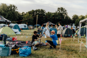 Zwei Männer beim Aufbau eines Zeltes auf dem Elbenwald Festival, umgeben von weiteren Zelten und Festivalbesuchern. Bunte Fähnchen schmücken den Campingbereich, während das Festivalleben im Hintergrund pulsiert.