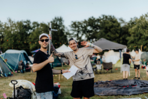 Zwei junge Männer auf dem Elbenwald Festival, einer hält ein Zeltgestänge, der andere winkt fröhlich. Im Hintergrund sind Zelte und Festivalbesucher zu sehen, die die entspannte Atmosphäre des Events genießen.
