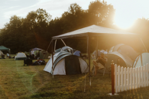 Zeltplatz beim Elbenwald Festival während des Sonnenuntergangs, mit verschiedenen Zelten im Hintergrund und einem Besucher, der entspannt sitzt. Die Atmosphäre ist entspannt und lädt zum Verweilen ein. Ideal für Nerds und Musikliebhaber.