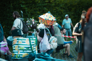 Eine Besucher*in des Elbenwald Festivals trägt einen bunten Hut und hält einen Blumenstrauß. Im Hintergrund sind andere Festivalteilnehmende zu sehen, die in entspannter Atmosphäre auf das Event warten. Die Szenerie spiegelt die kreative und lebendige Festivalatmosphäre wider.