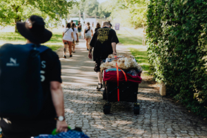 Person zieht einen Wagen mit Ausrüstung und Kostümen auf dem Weg zum Elbenwald Festival, umgeben von anderen Festivalbesuchern. Die Szene zeigt die lebendige Atmosphäre des Events inmitten der Natur.