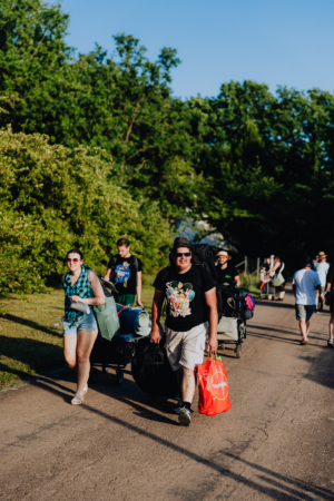 Festivalbesucher gehen mit Gepäck und Ausrüstung zum Elbenwald Festival, um die Mischung aus Musik und Nerdkultur zu genießen. Die Gruppe besteht aus mehreren Personen, die in einer sonnigen, grünen Umgebung unterwegs sind.
