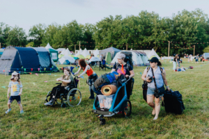 Besucher des Elbenwald Festivals spazieren über das Gelände, während sie Gepäck und Ausrüstung tragen. Im Hintergrund sind Zelte und andere Festivalgäste zu sehen, die die entspannte Atmosphäre des Events genießen.
