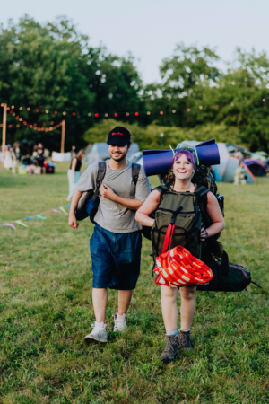 Zwei Festivalbesucher mit Rucksäcken und Campingausrüstung gehen über eine grüne Wiese beim Elbenwald Festival. Im Hintergrund sind Zelte und festliche Lichter sichtbar, die eine lebendige Atmosphäre schaffen.