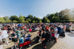 Menschenmenge wartet in der Schlange beim Elbenwald Festival, umgeben von Bäumen und einem klaren blauen Himmel. Teilnehmer tragen Rucksäcke und Gepäck, während sie sich auf das bevorstehende Festivalerlebnis vorbereiten.