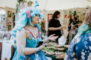 Artist Alley Eine cosplayerin mit blauen Haaren und maritimen Accessoires interagiert mit anderen Besuchern beim Elbenwald Festival, umgeben von kunstvollen Ständen und handgefertigten Produkten. Die Atmosphäre ist lebhaft und kreativ, typisch für ein nerdiges Event.