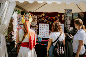 Artist Alley Besucherinnen des Elbenwald Festivals erkunden einen bunten Stand mit Merchandise, der mit kreativen Kunstwerken, Aufklebern und handgemachten Produkten geschmückt ist. Die Atmosphäre ist lebhaft und einladend, umgeben von festlicher Dekoration.