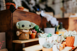 Artist Alley Handgefertigte, gehäkelte Figur eines beliebten Charakters in einem kreativen Verkaufsstand auf dem Elbenwald Festival. Umgeben von bunten Pflanzen in Töpfen, bietet der Stand eine einzigartige Mischung aus Kunst und Nerdkultur.
