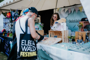 Artist Alley Besucher des Elbenwald Festivals stehen an einem Stand mit handgefertigten Accessoires und Hüten. Eine Verkäuferin lächelt freundlich, während sie die Produkte präsentiert. Der Stand ist dekoriert mit bunten Schildern und Blumen.