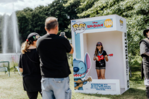 Ein Mädchen mit Ukulele posiert in einer Fotobox mit Stitch-Motiv auf dem Elbenwald Festival. Im Hintergrund sind Besucher und ein Springbrunnen zu sehen, während die Atmosphäre des Festivals lebendig und bunt ist.