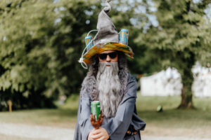 Ein verkleideter Zauberer mit grauem Bart und Sonnenbrille steht auf dem Elbenwald Festival. Er trägt einen auffälligen Hut mit Getränkedosen und hält eine Dose in der Hand. Der Hintergrund zeigt eine grüne Wiese und Festivalatmosphäre.