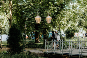 Zwei Besucher schlendern über eine Brücke im Elbenwald Festival, umgeben von grüner Natur und festlicher Beleuchtung. Die Atmosphäre ist lebhaft und einladend, ideal für Fans von Musik und Nerd-Kultur.