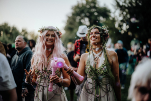 Zwei fröhliche Frauen in fantasievollen Kostümen und Blumenkränzen, die auf dem Elbenwald Festival stehen. Sie halten leuchtende Spielzeuge und umgeben von Seifenblasen, während die Festivalatmosphäre lebhaft und bunt ist.
