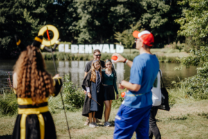 Besucher des Elbenwald Festivals posieren vor dem Festival-Schild am Ufer eines Sees. Im Vordergrund stehen zwei Mädchen in Kostümen, während ein weiterer Besucher mit einem Ball spielt. Eine lebendige Szene voller Spaß und kreativer Kostüme.