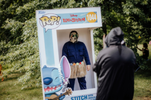 Ein Teilnehmer in einem Lilo & Stitch Kostüm steht in einer Fotobox beim Elbenwald Festival. Die Box zeigt die Funko Pop! Figur von Stitch mit einer Ukulele. Im Hintergrund sind weitere Festivalbesucher und eine grüne Umgebung zu sehen.
