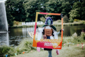 Cosplayer mit blauer Haut und lila Haaren posiert mit einem Rahmen beim Elbenwald Festival 2026, umgeben von einer malerischen Kulisse am Wasser. Im Hintergrund ist das Festival-Logo zu sehen. Bunte Fahnen und ein Wasserfall ergänzen die festliche Atmosphäre.