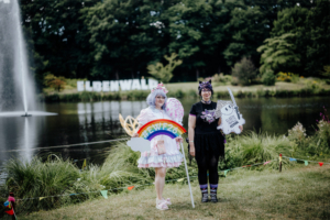 Zwei Teilnehmerinnen des Elbenwald Festivals posieren am Ufer eines Teichs, umgeben von bunten Fähnchen. Eine trägt ein pastellfarbenes Kostüm mit Regenbogen und die andere ein schwarzes Outfit mit Katzenohren. Im Hintergrund ist das Festival-Logo sichtbar.