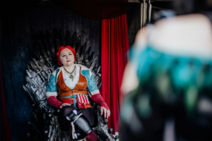 Ein Cosplayer sitzt auf einem Thron aus Schwertern, gekleidet in ein fantasievolles Kostüm mit rotem Haar und einer auffälligen Halskette. Im Hintergrund sind rote Vorhänge sichtbar, die eine festliche Atmosphäre beim Elbenwald Festival schaffen.