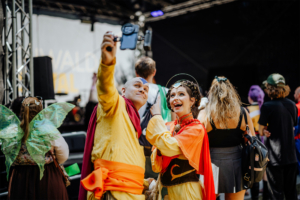 Zwei verkleidete Festivalbesucher machen ein Selfie beim Elbenwald Festival. Im Hintergrund sind weitere Teilnehmer in bunten Kostümen zu sehen, die die lebendige Atmosphäre des Events widerspiegeln.