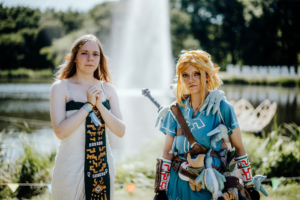 Zwei Cosplayerinnen auf dem Elbenwald Festival, eine verkleidet als Link aus "The Legend of Zelda" und die andere in traditioneller Kleidung. Im Hintergrund sprudelt ein Brunnen, umgeben von grünem Gras und Bäumen, was die festliche Atmosphäre unterstreicht.
