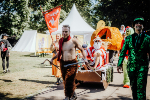 Bunte Parade beim Elbenwald Festival mit verkleideten Teilnehmern, darunter ein Weihnachtsmann in einem Schlitten, ein Grinch und weitere fantasievolle Kostüme. Festliche Atmosphäre und kreative Darstellungen auf dem Festivalgelände.