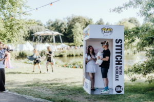 Zwei Festivalbesucher posieren in einer Fotobox mit dem Thema "Stitch mit Ukulele" beim Elbenwald Festival. Im Hintergrund sind Zelte und andere Teilnehmer zu sehen, die die fröhliche Atmosphäre des Events genießen.