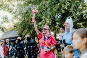 Eine fröhliche Festivalbesucher*in mit pinken Haaren und Sonnenbrille, die eine Seifenblasenmaschine hält und in einem bunten Kostüm gekleidet ist. Im Hintergrund sind weitere Teilnehmer*innen in verschiedenen Outfits zu sehen, die die lebhafte Atmosphäre des Elbenwald Festivals genießen.
