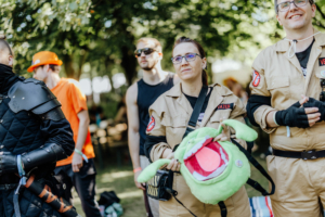 Teilnehmer in Kostümen beim Elbenwald Festival, darunter eine Frau in einem Ghostbusters-Outfit mit einem grünen Plüschmonster. Im Hintergrund sind weitere Festivalbesucher zu sehen, die eine entspannte Atmosphäre im Freien genießen.