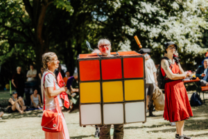 Bunt kostümierte Festivalbesucher auf dem Elbenwald Festival, darunter ein Mann in einem Rubik's Cube-Kostüm, umgeben von fröhlichen Teilnehmern in einem grünen Park. Die Atmosphäre ist lebhaft und feierlich, perfekt für Fans von Musik und Popkultur.
