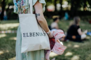 Eine Person hält eine Stofftasche mit dem Aufdruck "ELBENWALD" und einem bunten Tuch in der Hand, während im Hintergrund andere Festivalbesucher auf dem Rasen sitzen. Die Szene fängt die entspannte Atmosphäre des Elbenwald Festivals ein.