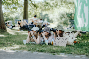 Drei Cosplayer mit Kameras liegen auf dem Gras beim Elbenwald Festival. Sie fotografieren Passanten und genießen die entspannte Festivalatmosphäre. Im Hintergrund sind andere Besucher und ein Schild mit der Aufschrift „COSPLAY SAFARI“ zu sehen.