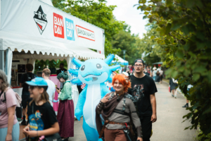Buntes Treiben beim Elbenwald Festival: Besucher in kreativen Kostümen, darunter ein großer blauer Axolotl, schlendern an einem Stand von Bandai vorbei, umgeben von grüner Natur und fröhlicher Festivalatmosphäre.