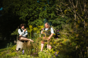 Zwei Männer im historischen Kostüm sitzen entspannt in einer blühenden Gartenlandschaft während des Elbenwald Festivals. Einer raucht eine Pfeife, während der andere lässig auf einem Baumstumpf sitzt, umgeben von bunten Blumen und grünen Pflanzen.