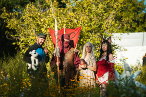 Vier kostümierte Teilnehmer des Elbenwald Festivals posieren in einer bunten, blühenden Landschaft. Die Gruppe zeigt eine Vielfalt an Charakteren, darunter ein Krieger mit einer Fahne, eine Drachenmutter und eine Figur mit Katzenohren, umgeben von Pflanzen.
